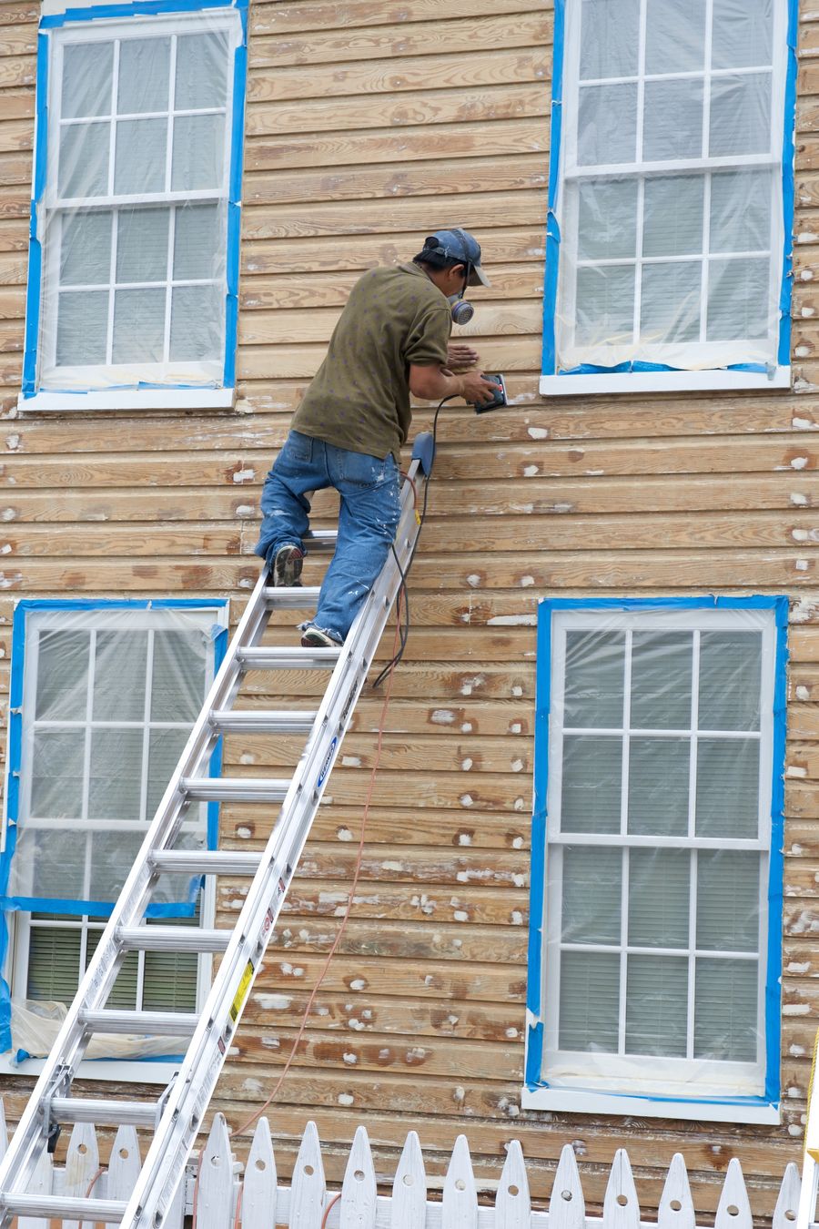 Painter on an extension ladder sanding weathered clapboard siding, with the windows masked in plastic before repainting
