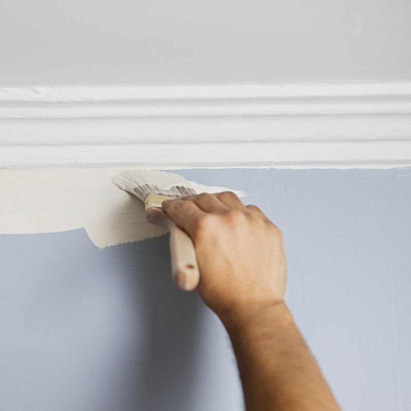 Hand brushing white paint onto a blue-gray wall below white molding